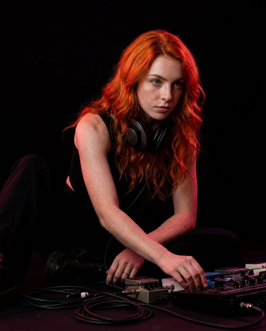 Celeste sitting on the studio floor, headphones round her neck, surrounded by cables and pedals.